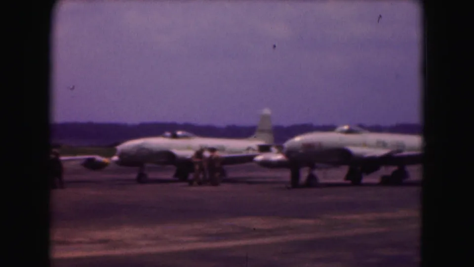 Uniformed personnel appear near two parked military jets during routine ground activity in this 1975 8mm air base scene.