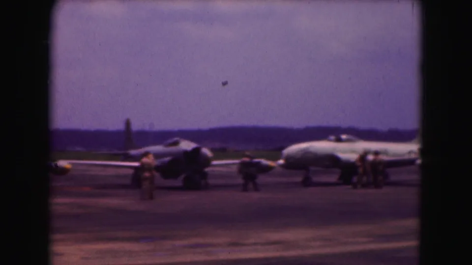 Several people move around parked military jets on the tarmac, with a low horizon and purple-shifted 8mm color.