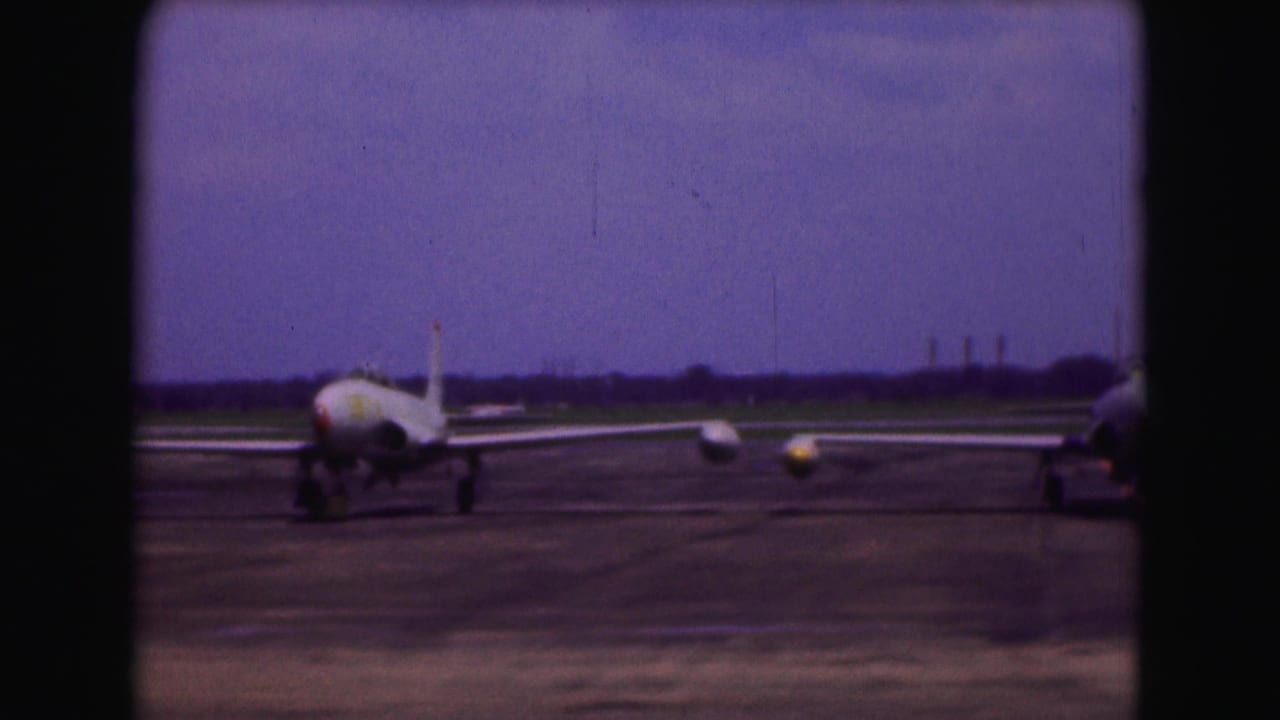 1975 Washington, DC home-movie frame showing a military jet aircraft on a runway under a purple-tinged sky.