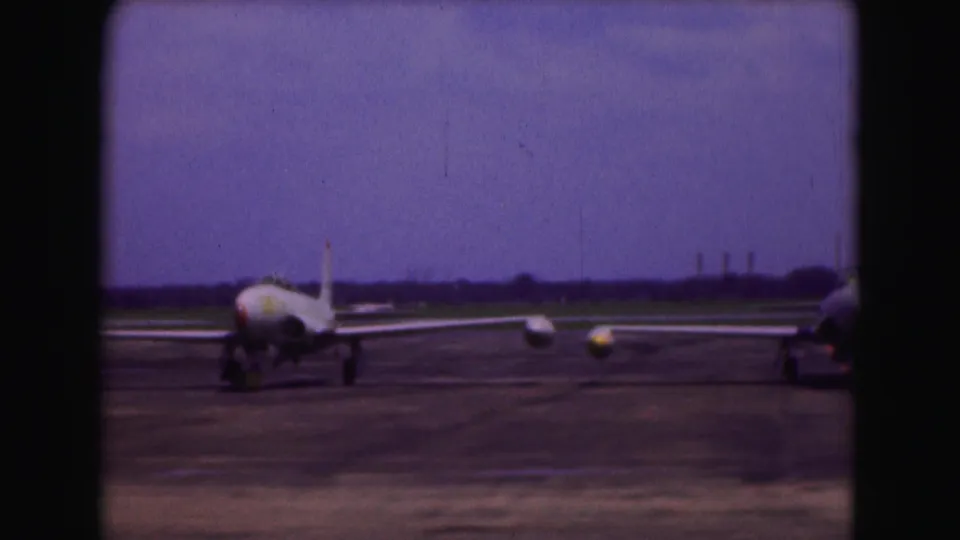 1975 Washington, DC home-movie frame showing a military jet aircraft on a runway under a purple-tinged sky.