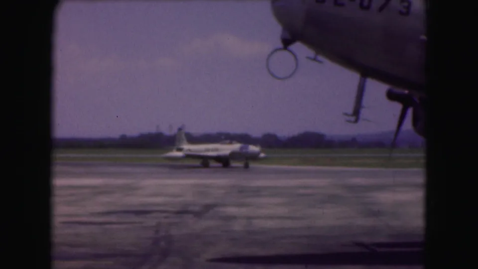 A 1975 airport frame shows the taxiing jet beyond the tarmac while the nose or fuselage of another aircraft enters the upper right foreground.