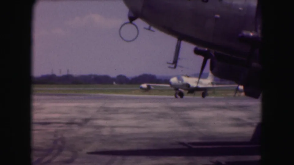 The jet turns across the 1975 airfield scene, partly framed by the large parked aircraft in the foreground.