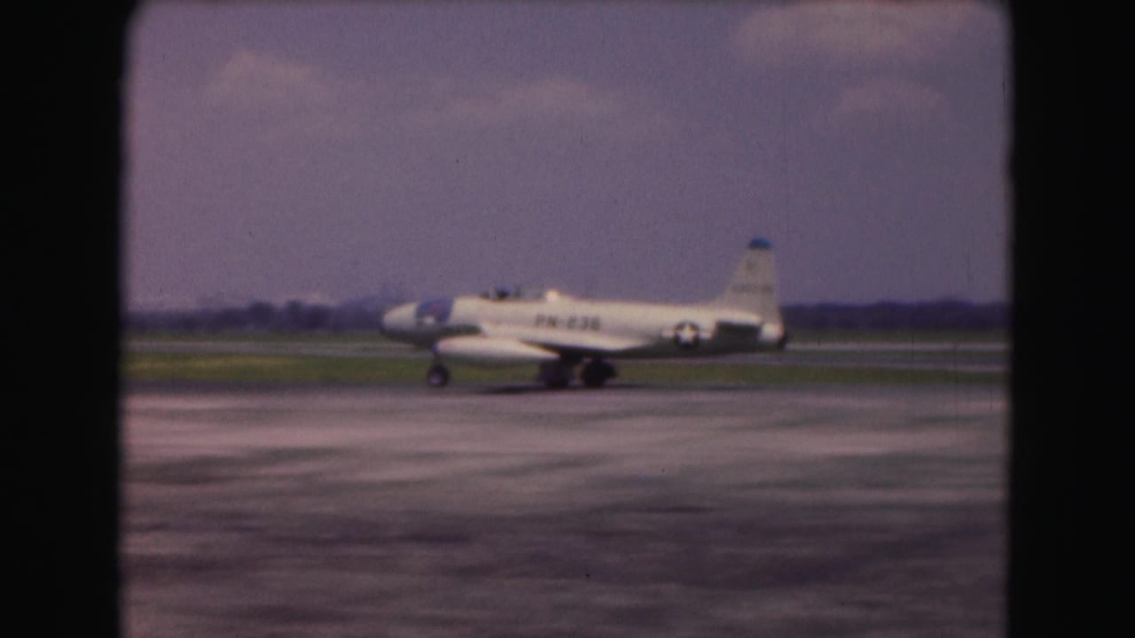 1975 Washington DC 8mm frame showing a small white military-marked aircraft taxiing across runway pavement under a hazy sky.