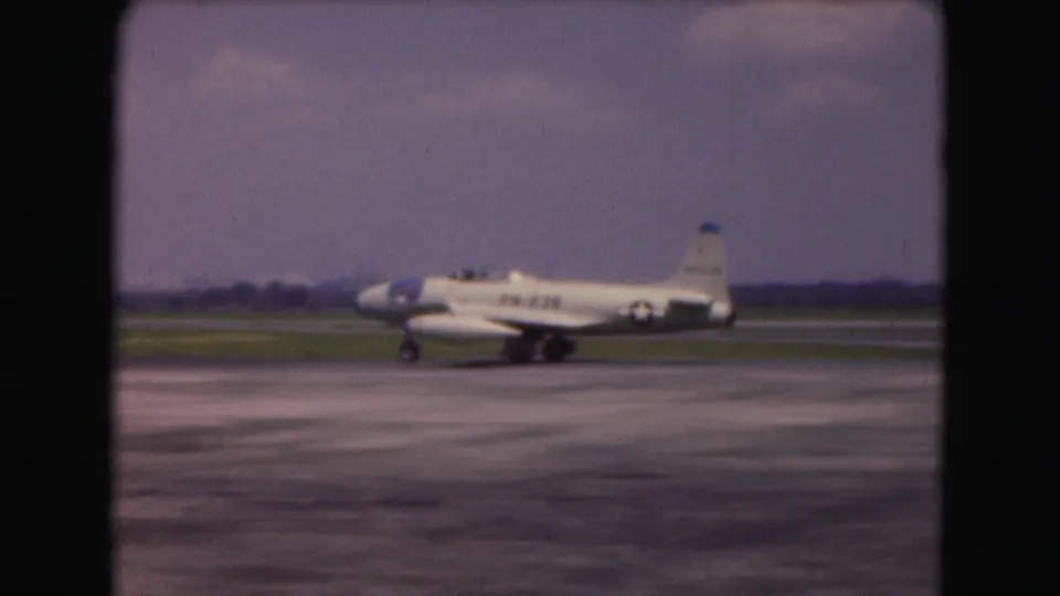1975 Washington DC 8mm frame showing a small white military-marked aircraft taxiing across runway pavement under a hazy sky.