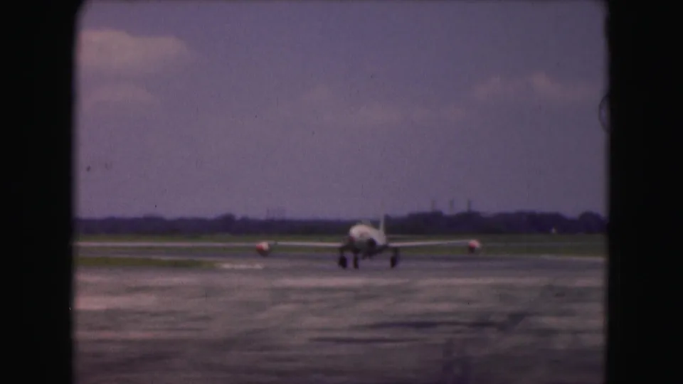 1975 Washington DC 8mm frame with the aircraft farther along the runway, viewed front-on against grass, treeline, and hazy sky.