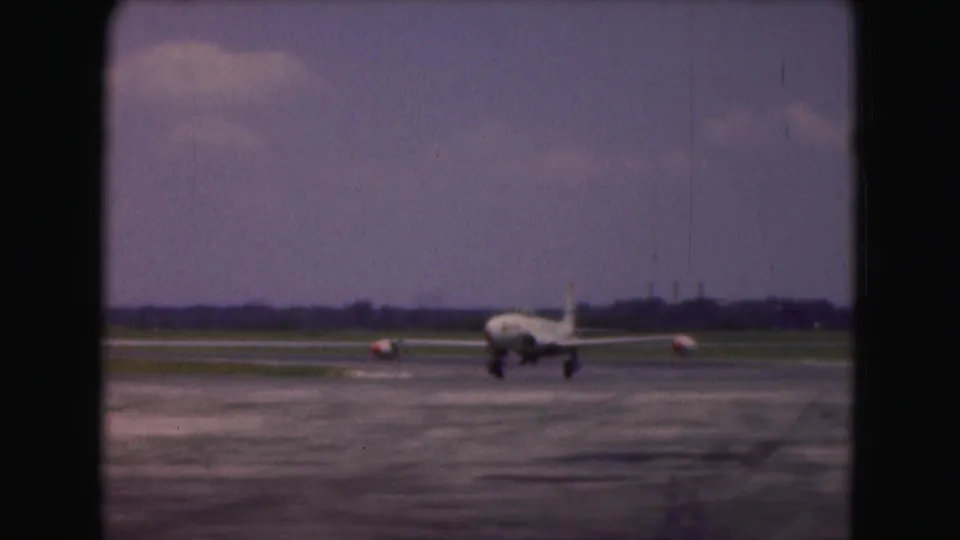 1975 Washington DC home-movie frame showing a white USAF jet approaching across runway pavement beneath a cloudy sky.