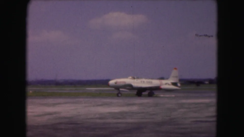 1975 Washington DC home-movie frame showing the side of a white USAF jet taxiing past grassy airfield margins.