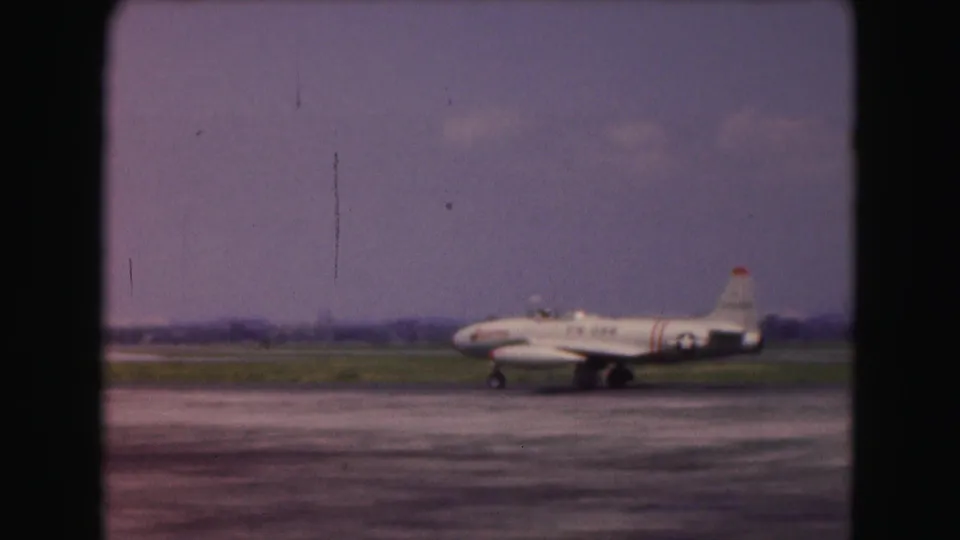 Soft 8mm frame of a white jet rolling across an airport runway, with muted sky and distant landscape behind it.