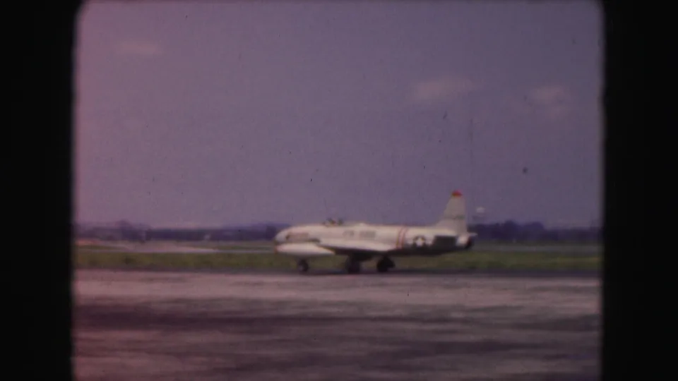 1975 8mm airport footage shows the white jet farther along the tarmac beneath a hazy purple-blue sky.