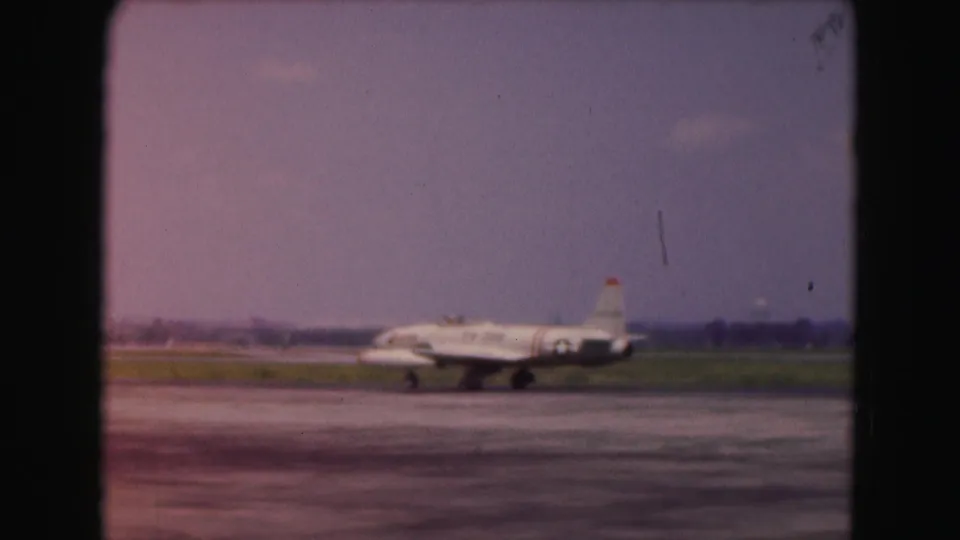 1975 Washington, DC-area home-movie frame showing a small jet aircraft on an airfield, with grass and distant structures behind it.