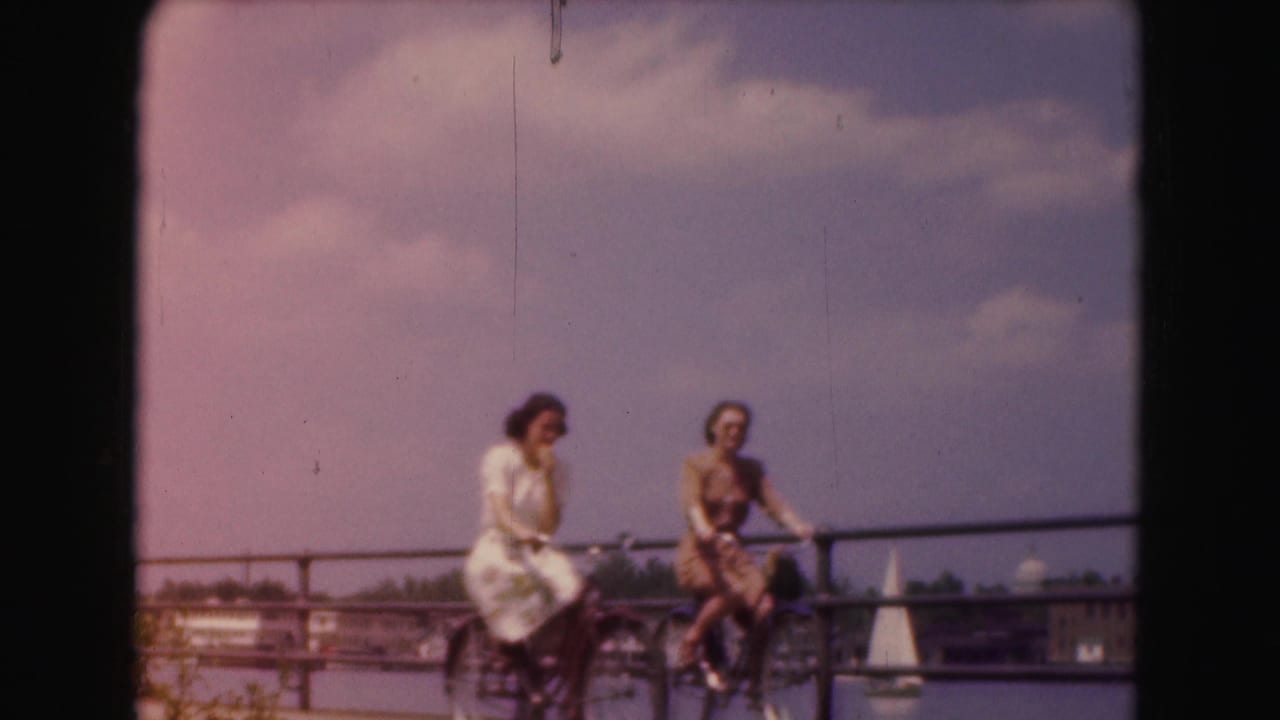 Two women in 1970s attire pause with bicycles along a Washington, DC waterfront promenade, with water and a sailboat in the distance.
