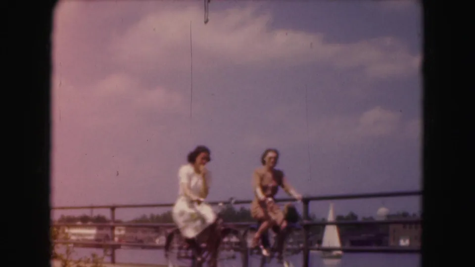 Two women in 1970s attire pause with bicycles along a Washington, DC waterfront promenade, with water and a sailboat in the distance.