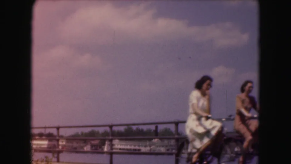 1975 Washington, DC home-movie frame of two women riding or pausing with bicycles along a waterfront railing, with water and buildings beyond.
