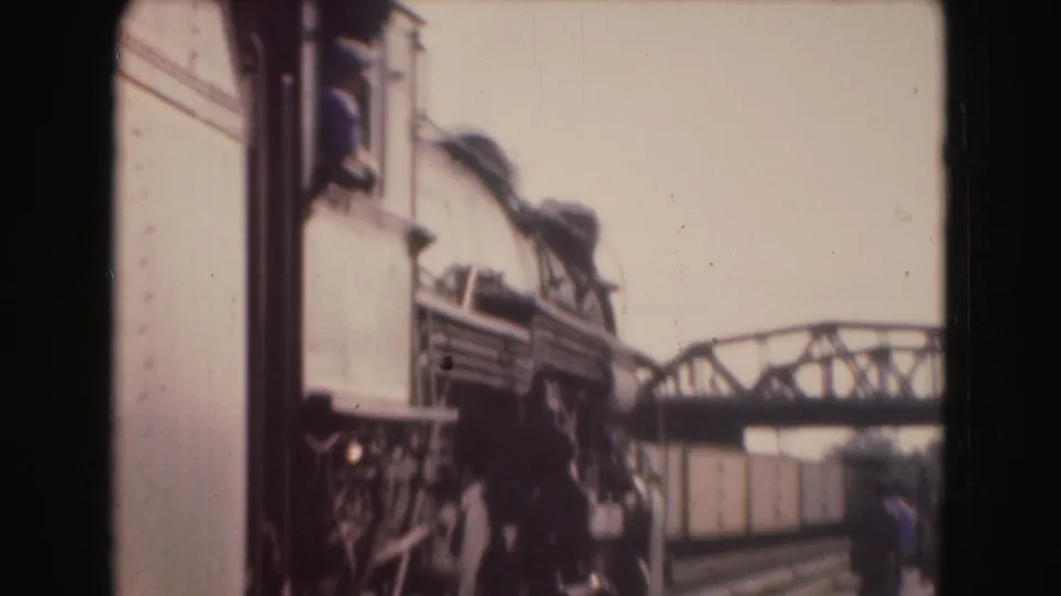 Soft 8mm frame showing a rail-side or train-side view with metal structure, smoke or shadowed machinery, and a bridge in the background.