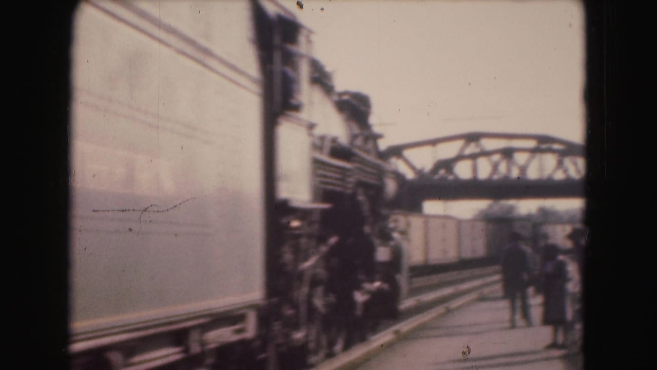 Train passing a station platform with a few people standing near the tracks.