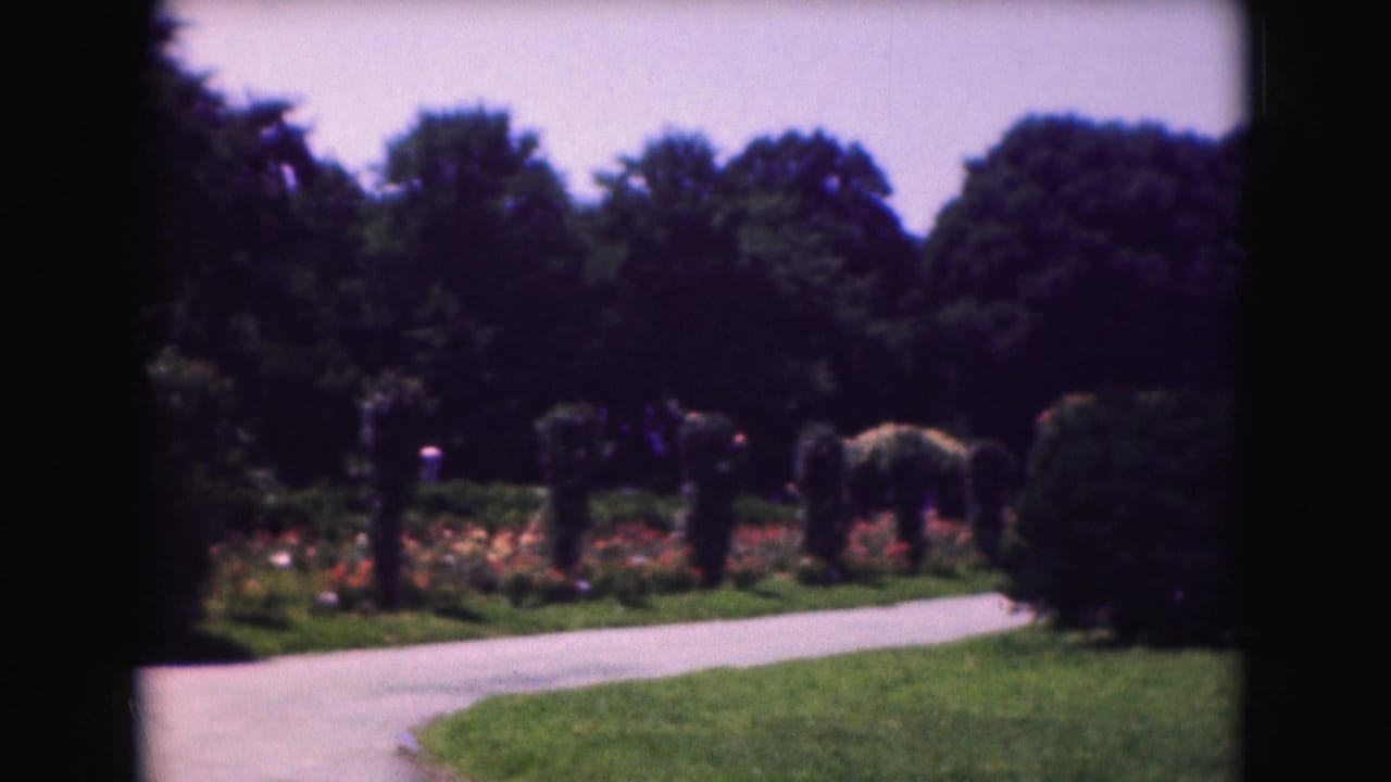 1975 Washington, DC garden view with a curving path, flower beds, dense trees, lawn, and distant visitors.