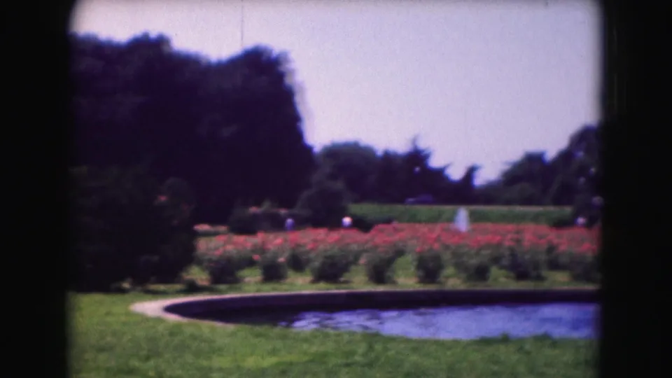 1975 Washington, DC garden view with a pond, red flower beds, trees, hedge line, distant visitors, and a small fountain.