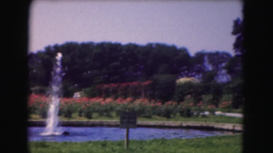 1975 Washington DC 8mm frame showing a pond fountain beside red flower beds, a paved path, green grass, trees, and a small sign.