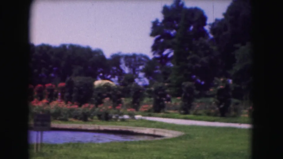 1975 Washington DC visitor-shot garden view with a pond edge, curved path, red flower beds, open lawn, mature trees, and a sign in the foreground.