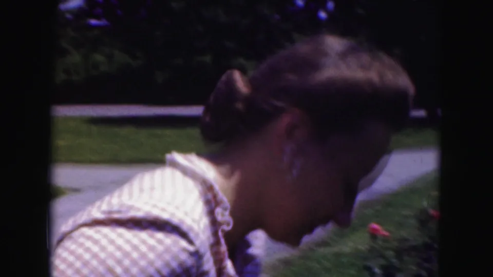 Young woman leans down toward garden flowers in a 1975 Washington, DC home-movie frame.