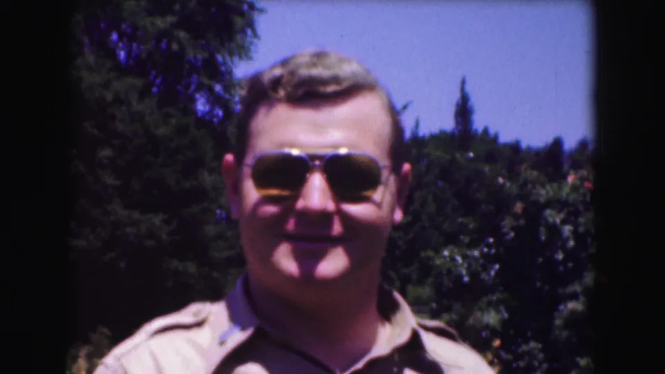 A man wearing aviator sunglasses smiles faintly toward the camera, framed by trees and blue sky in the Washington DC home movie.