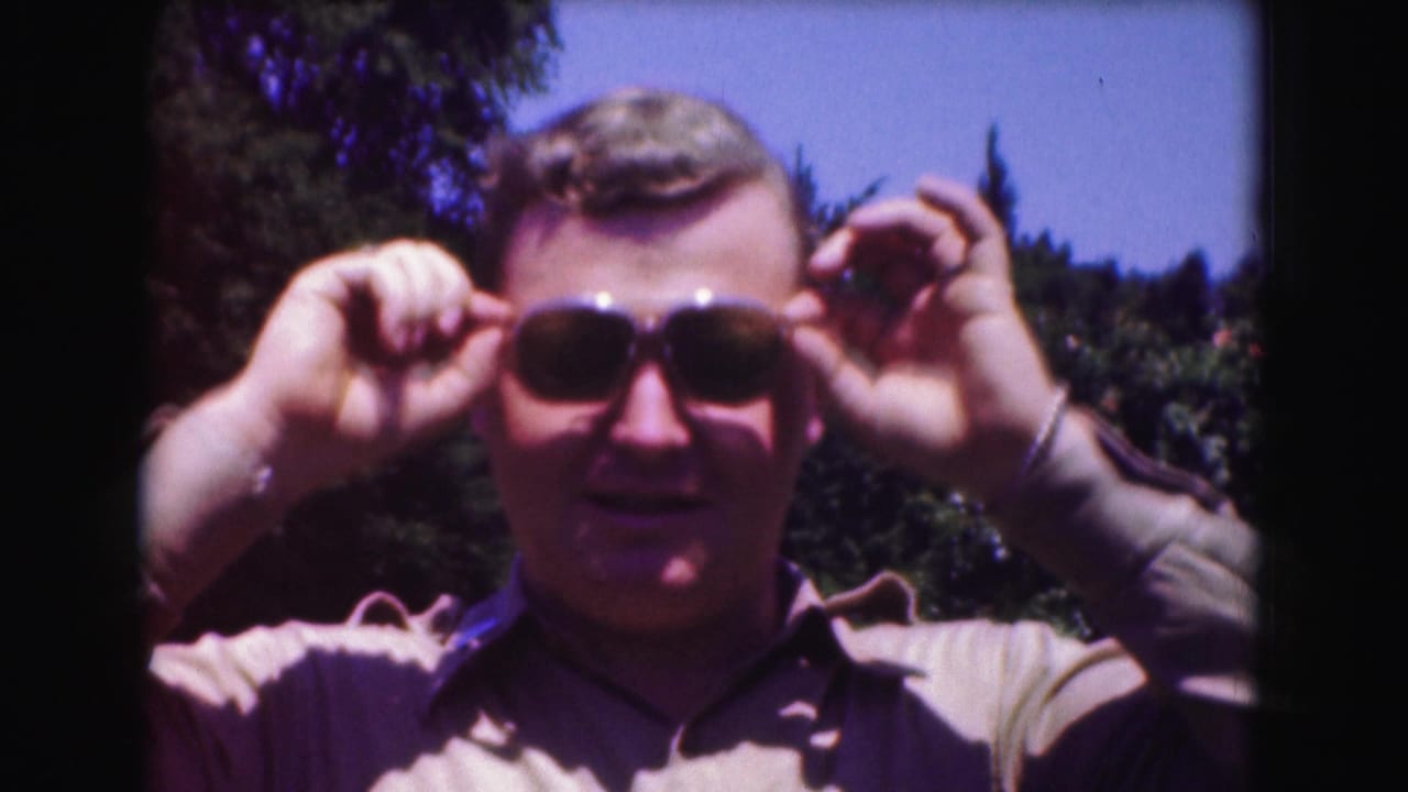 The man lifts both hands to his aviator sunglasses while standing against trees and blue sky in 1975 Washington, DC footage.
