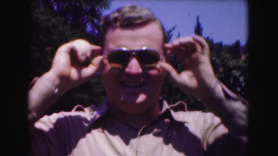 A smiling adult man holds both sides of his aviator sunglasses in a sunlit 1975 Washington, DC home-movie frame.