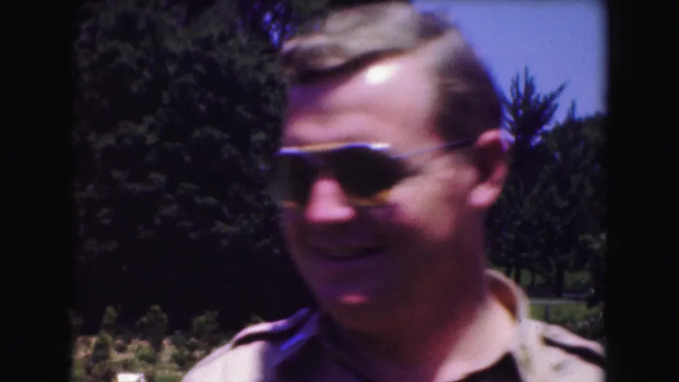 The man turns slightly to the side, still wearing aviator sunglasses, with green trees behind him in the 1975 home-movie shot.