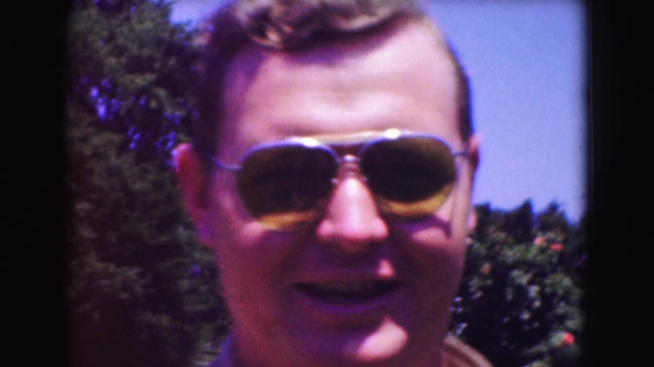 Man wearing aviator sunglasses smiles outdoors beside greenery.