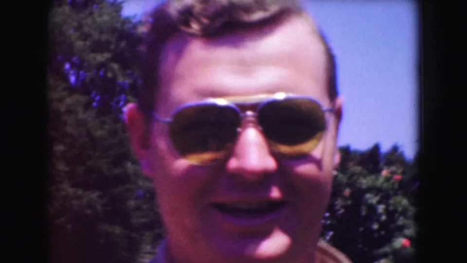 In 1975 Washington DC home-movie footage, a man in aviator sunglasses faces the camera near garden foliage.
