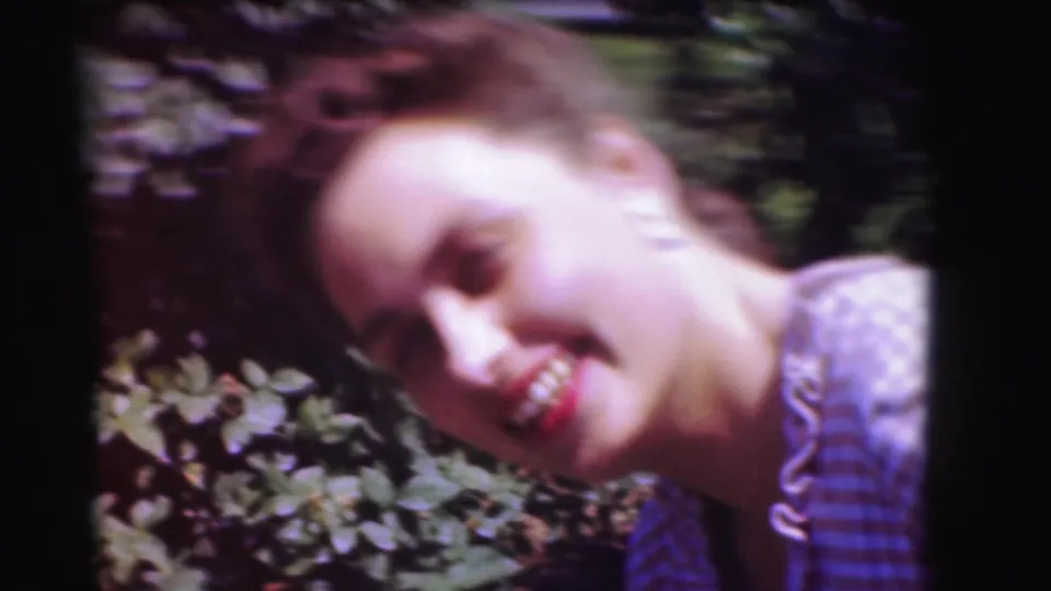 A smiling woman in a blue checked dress leans toward the camera beside garden foliage in 1975 Washington, DC home-movie footage.