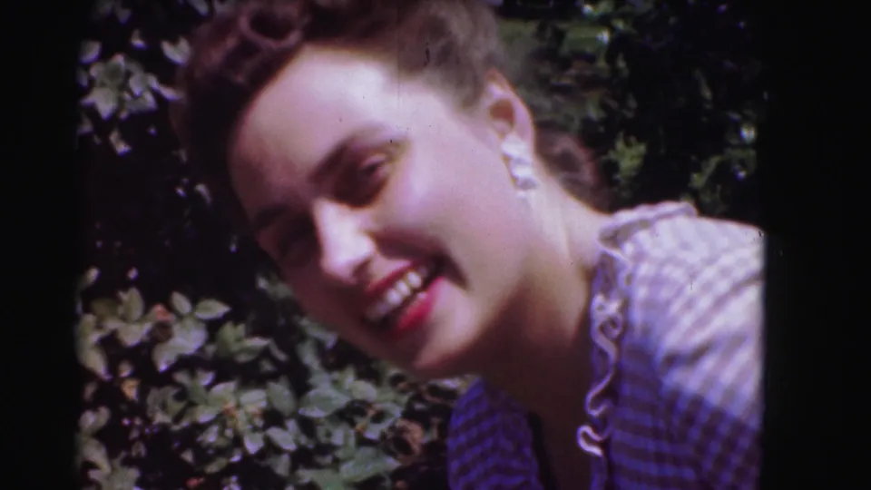 A woman smiles outdoors beside foliage in 1975 Washington, DC home-movie footage, wearing a checkered ruffled dress and white earrings.