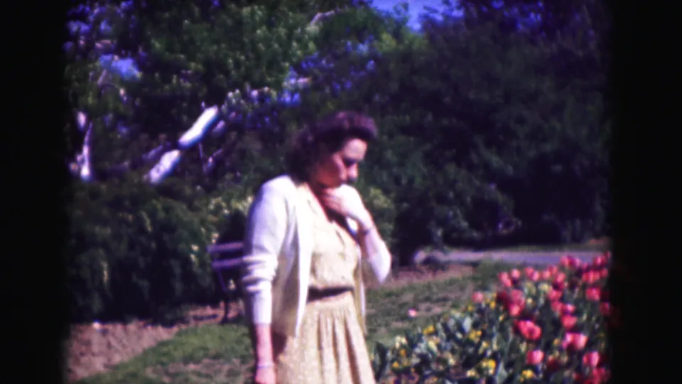 1975 Washington DC frame showing a woman in a pale dress and cardigan beside tulips, with a path, bench, and trees behind her.