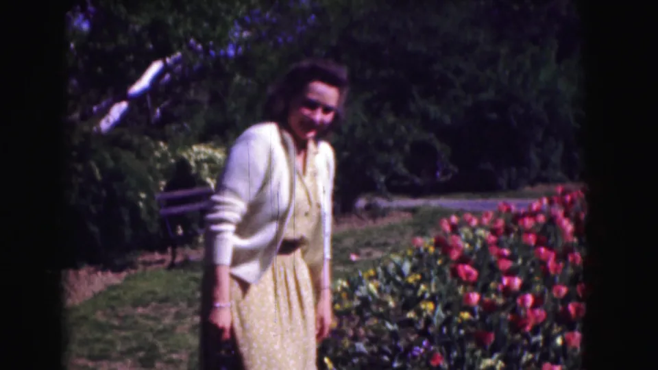 The woman smiles toward the camera beside red tulips, with trees and a bench in the background.