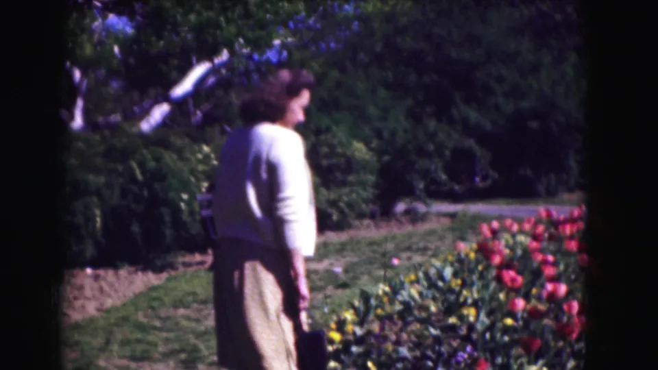 Seen from behind, the woman walks beside tulips and a lawn, with trees and shrubs filling the garden background.