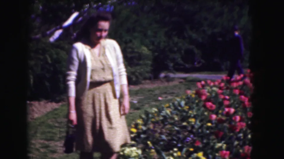 The woman faces the camera near tulip beds while a distant visitor walks along the garden path in Washington DC footage from 1975.