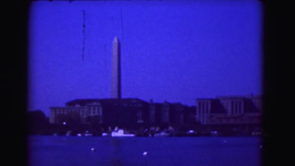 Blue-tinted 1975 8mm view across water toward the Washington Monument in Washington, DC, with buildings and small boats visible.