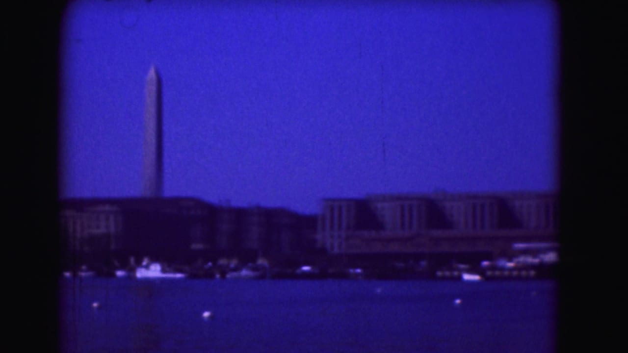 1975 Washington, DC home-movie frame showing the Washington Monument, Potomac River, and distant waterfront buildings under a deep blue sky.