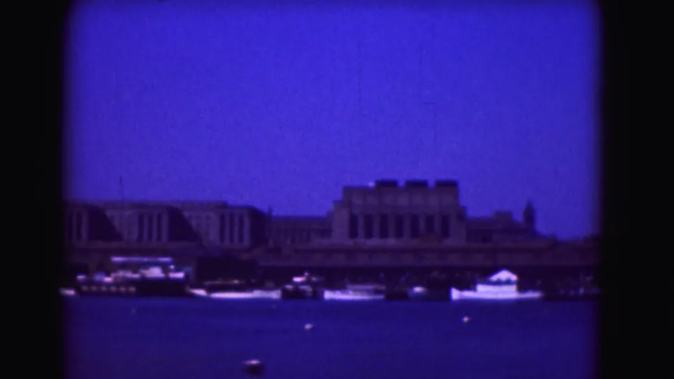 1975 Washington, DC home-movie frame showing river boats, waterfront structures, and a distant spire as the camera continues across the shoreline.