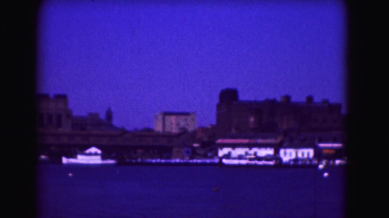 1975 8mm frame of a distant waterfront shoreline, with low buildings and boats across dark water under a blue-purple evening sky.