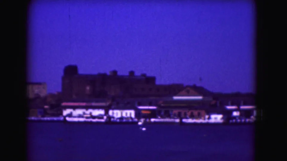 1975 8mm frame looking across a harbor toward low waterfront buildings and small boats, with a strong blue-purple film cast.