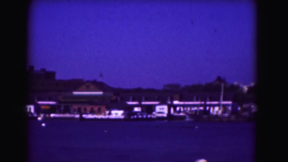 1975 8mm frame of a harbor shoreline with low buildings, boats, and a pier or marina area viewed from across the water.