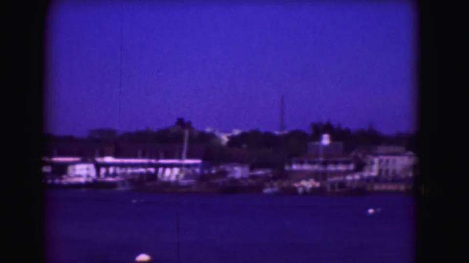 1975 8mm frame showing boats and shoreline structures across harbor water, with a tall mast or pole visible against the blue sky.