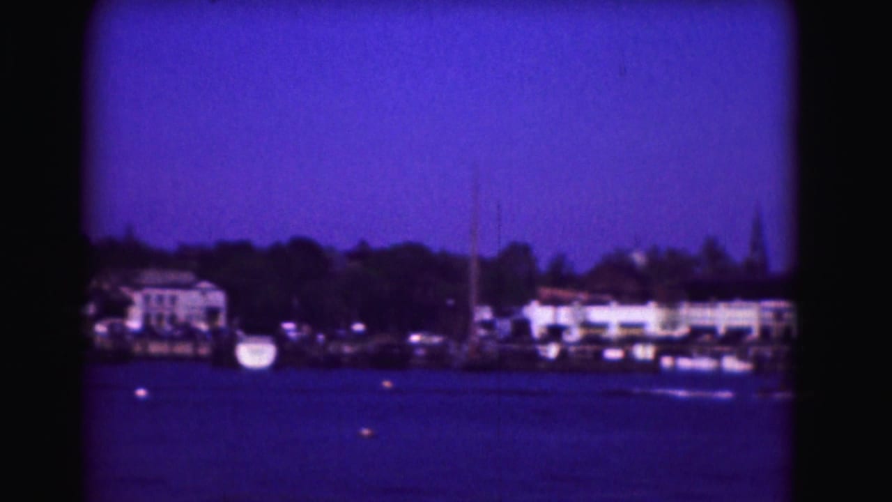 Distant shoreline buildings and trees across blue water with soft 8mm framing.