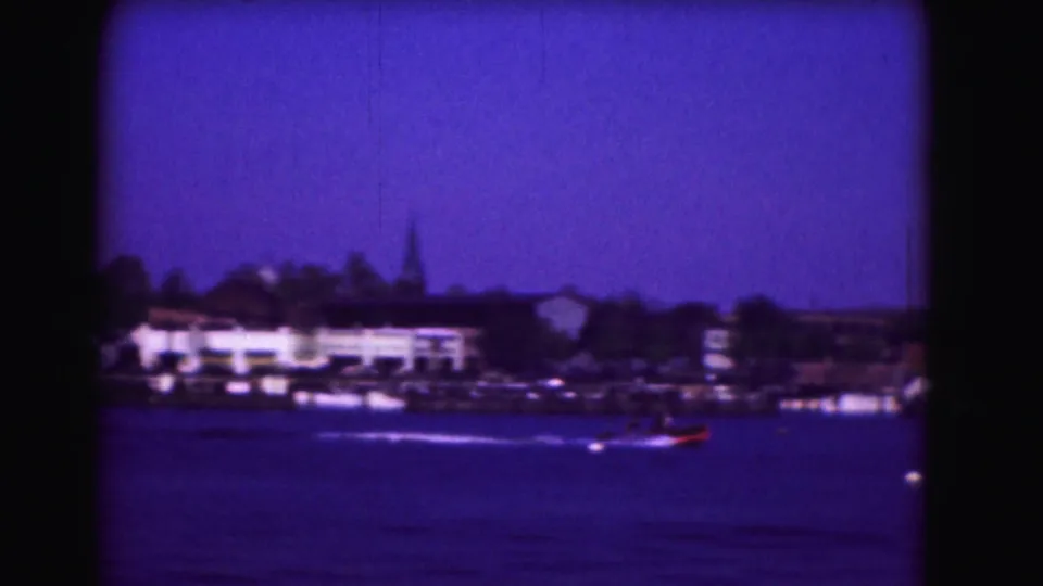 A small motorboat moves across blue water near a likely Washington, DC shoreline, with buildings and trees behind it.