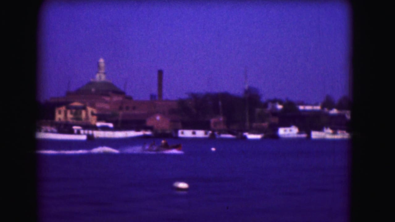 Speedboat on blue water with a white wake, docked boats, buildings, and a domed structure behind.