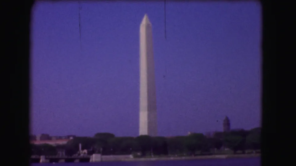 1975 Washington DC home-movie frame showing the Washington Monument across the Potomac with trees, shoreline, and dark 8mm frame edges.