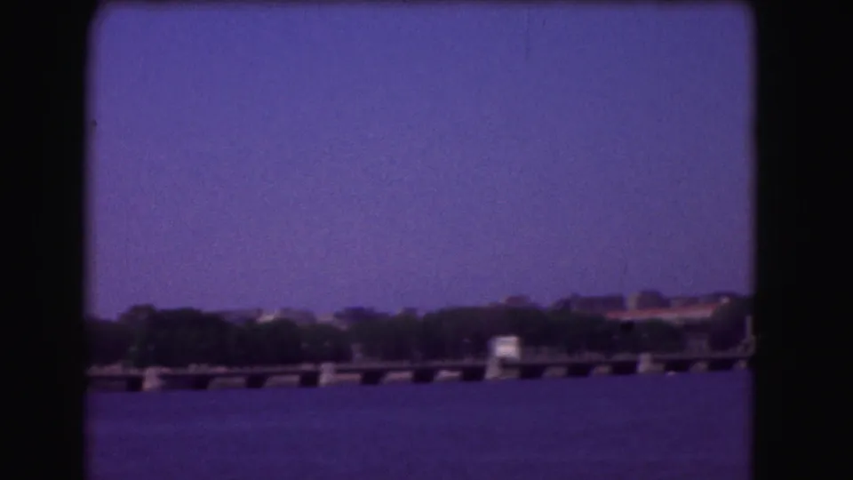 1975 visitor-shot frame across the Potomac, showing open water, a low waterfront structure, trees, and distant buildings under a blue-purple sky.