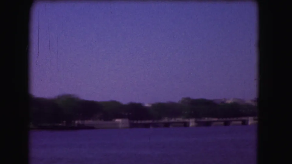 Washington DC home-movie frame with the Potomac in the foreground, a low pier or bridge, tree line, and distant buildings softened by 8mm color shift.