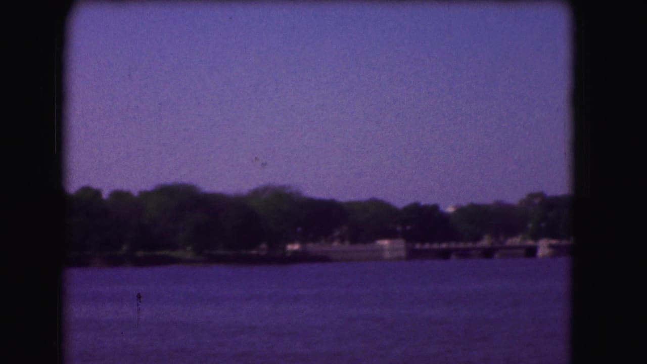 1975 Washington, DC home-movie frame showing open water and a distant wooded shoreline from a boat-like viewpoint.